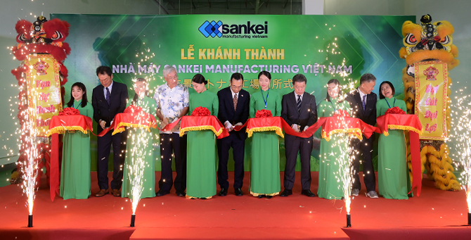 アオザイを着た社員と御来賓の皆様によるテープカットでは、幅広の布をはさみで切る。両側には獅子舞の獅子。手前は花火が噴き上がっている。